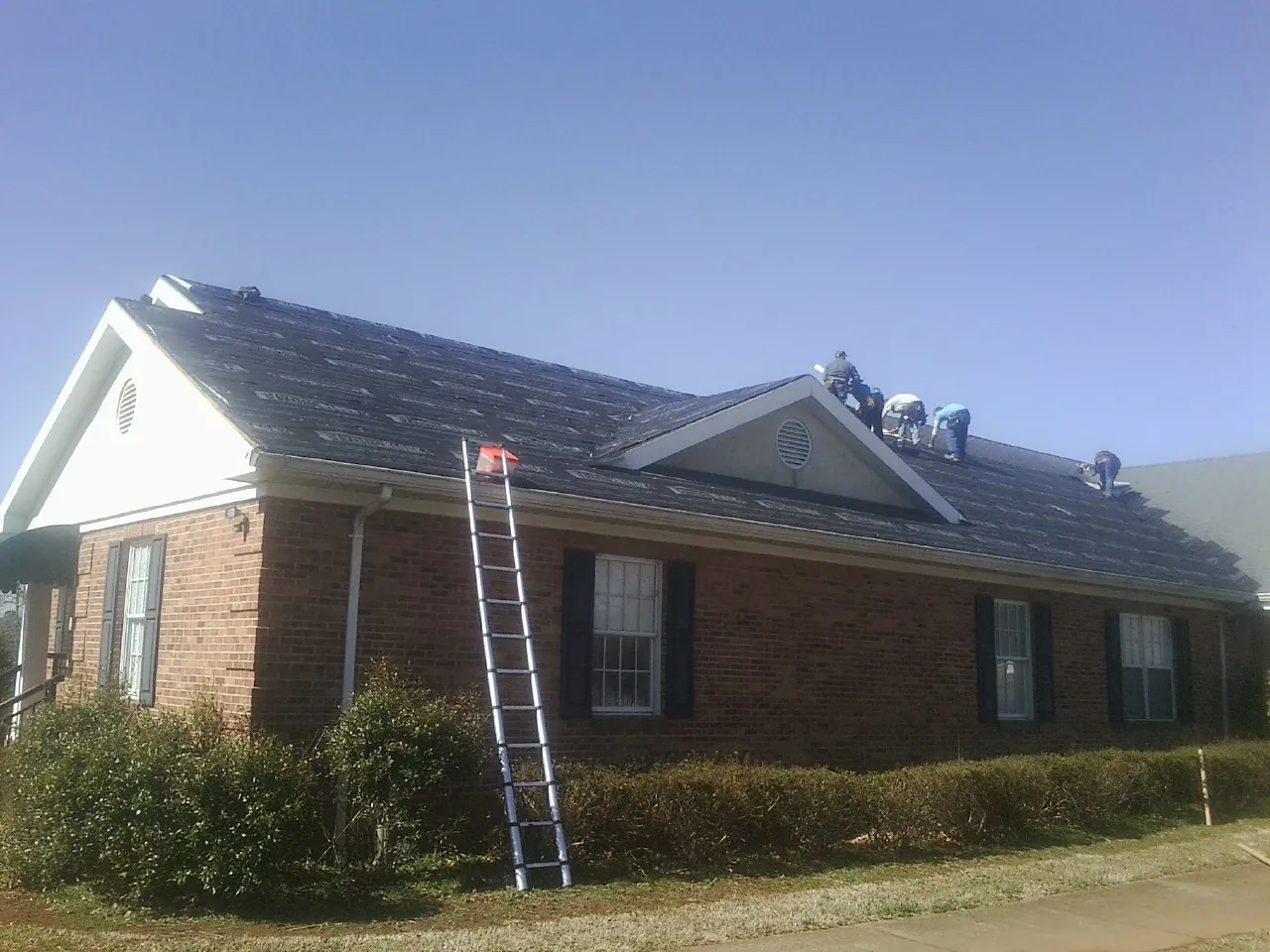 Crew tearing shingles off a brick ranch roof with a ladder set — Emergency Roof Repair work by our crew in Los Molinos
