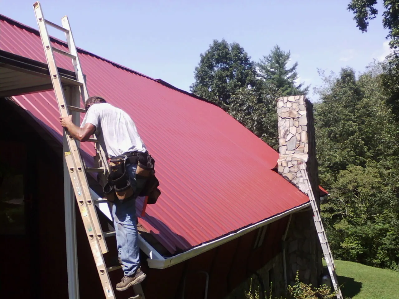 Roofer on a ladder beside a red metal roof and stone chimney — Soffit & Fascia Repair by Roofing Brothers in Los Molinos